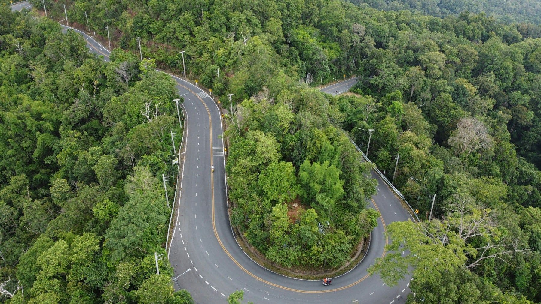 Eine Haarnadelkurve an einer bewaldeten Bergstraße aus der Luft, mit einem Motorrad, das außen um die Kurve fährt