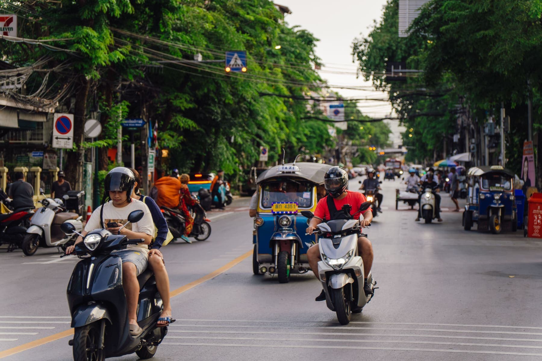 Eine baumgesäumte Straße mit Motorrollern und einem Tuk-Tuk zwischen parkenden Fahrzeugen