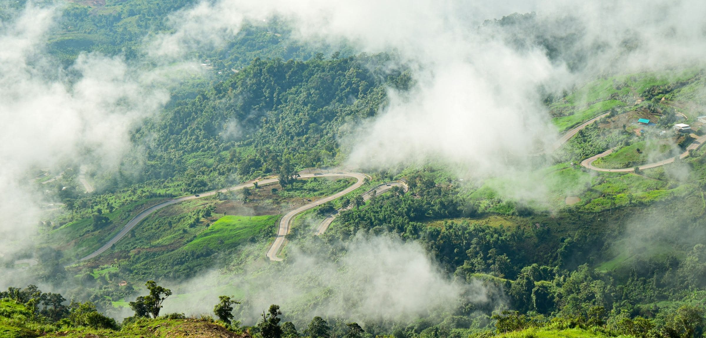 Eine Straße, die sich in Serpentinen durch steile bewaldete Hügel in Thailand windet, mit tiefen Wolken im Tal darunter